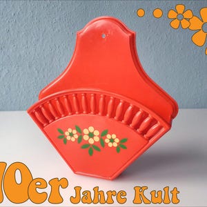 Peut inclure: Une poche murale orange vif, avec une base en forme d'éventail ornée de motifs floraux blancs et jaunes. Le texte "70er Jahre Kult" est affiché en orange. Cet article est probablement des années 1970.