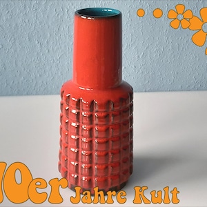 May include: A red ceramic vase with a textured, grid-like pattern on the lower portion and a smooth, cylindrical top. The interior is teal. The image includes the text "70er Jahre Kult" in orange, and orange flower graphics.