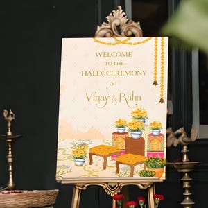 May include: A welcome sign for a Haldi ceremony, displayed on a gold easel. The sign features floral illustrations and the names "Vinay & Raha." The scene includes white vases, flowers, and decorative lamps, creating a festive atmosphere.