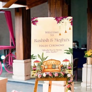 Può includere: Un cavalletto di legno espone un cartello con la scritta "Welcome to Kashish & Megha's Haldi Ceremony". Il cartello presenta un'illustrazione ad acquerello di un divano, ombrelli e composizioni floreali. La scena è decorata con fiori in vasi bianchi.