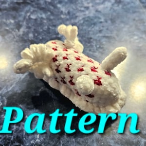 May include: A white crocheted plush toy with red accents in the shape of a sea slug. The toy has a textured surface and is lying on a blue and white tiled surface. The word "Pattern" is written in blue text below the toy.