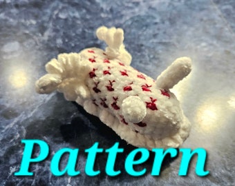 May include: A white crocheted plush toy with red accents in the shape of a sea slug. The toy has a textured surface and is lying on a blue and white tiled surface. The word "Pattern" is written in blue text below the toy.