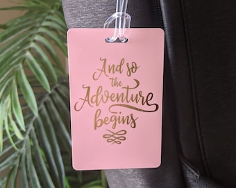 Op de afbeelding: Roze bagagelabel met gouden opschrift "And so the adventure begins".