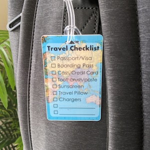 Op de afbeelding: Een rechthoekig reischecklistlabel met een wereldkaartontwerp en een doorzichtige plastic lus. Het label bevat reisbenodigdheden zoals paspoort, instapkaart en opladers. Het label is bevestigd aan een grijze rugzak.