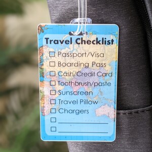 Op de afbeelding: Een rechthoekig reislabel met een doorzichtige plastic lus bevestigd aan een zwarte tas. Het label heeft een blauwe achtergrond met een wereldkaartontwerp en de woorden "Travel Checklist". De checklist bevat items zoals paspoort, boarding pass en opladers.