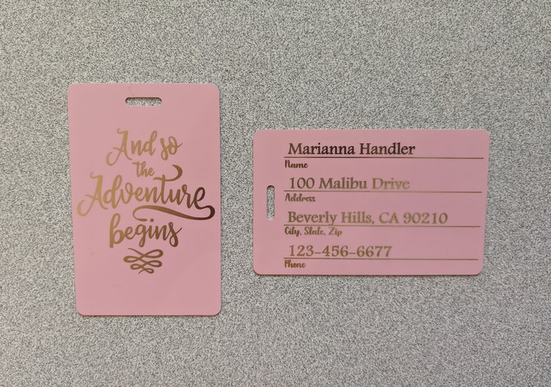 May include: Pink luggage tag with gold lettering that says "And so the Adventure begins". A second pink luggage tag with gold lettering has the text "Marianna Handler", "Name", "100 Malibu Drive", "Address", "Beverly Hills, CA 90210", "City, State, Zip", "123-456-6677", "Phone".