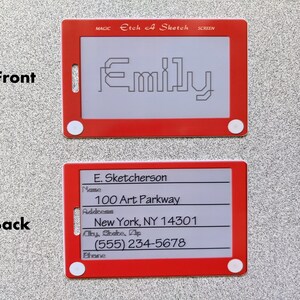 May include: Red plastic Etch A Sketch toy with a white screen. The screen shows the name "Emily" drawn in black lines. The back of the toy has a white screen with black text that reads "E. Sketcherson", "Name", "100 Art Parkway", "Address", "New York, NY 14301", "City, State, Zip", "(555) 234-5678", "Phone".