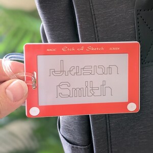 May include: Red Etch A Sketch luggage tag with the name "Jason Smith" drawn on the screen. The tag has a clear plastic loop for attaching to a bag.