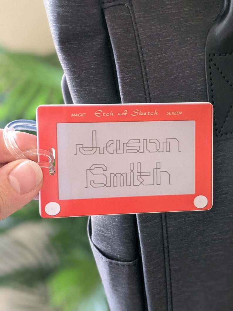 May include: Red Etch A Sketch luggage tag with the name "Jason Smith" drawn on the screen. The tag has a clear plastic loop for attaching to a bag.