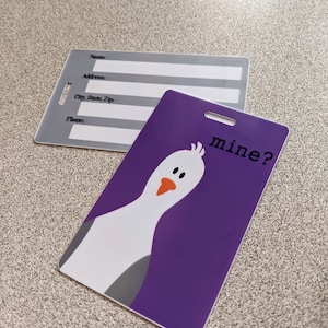 May include: Purple luggage tag with a white cartoon bird and the text "mine?" in black.