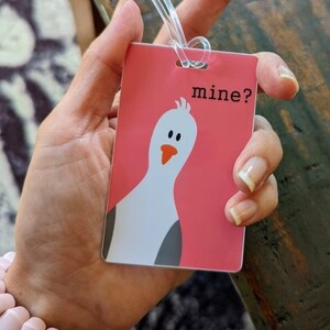 May include: Pink luggage tag with a cartoon white bird and the text "mine?" printed on it.