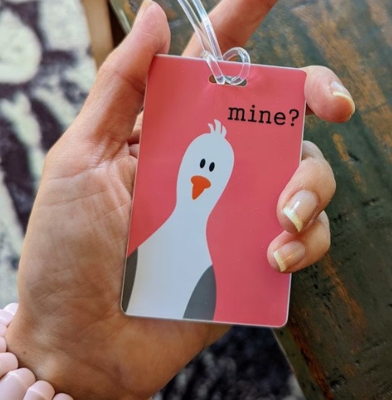 May include: Pink luggage tag with a cartoon white bird and the text "mine?" printed on it.