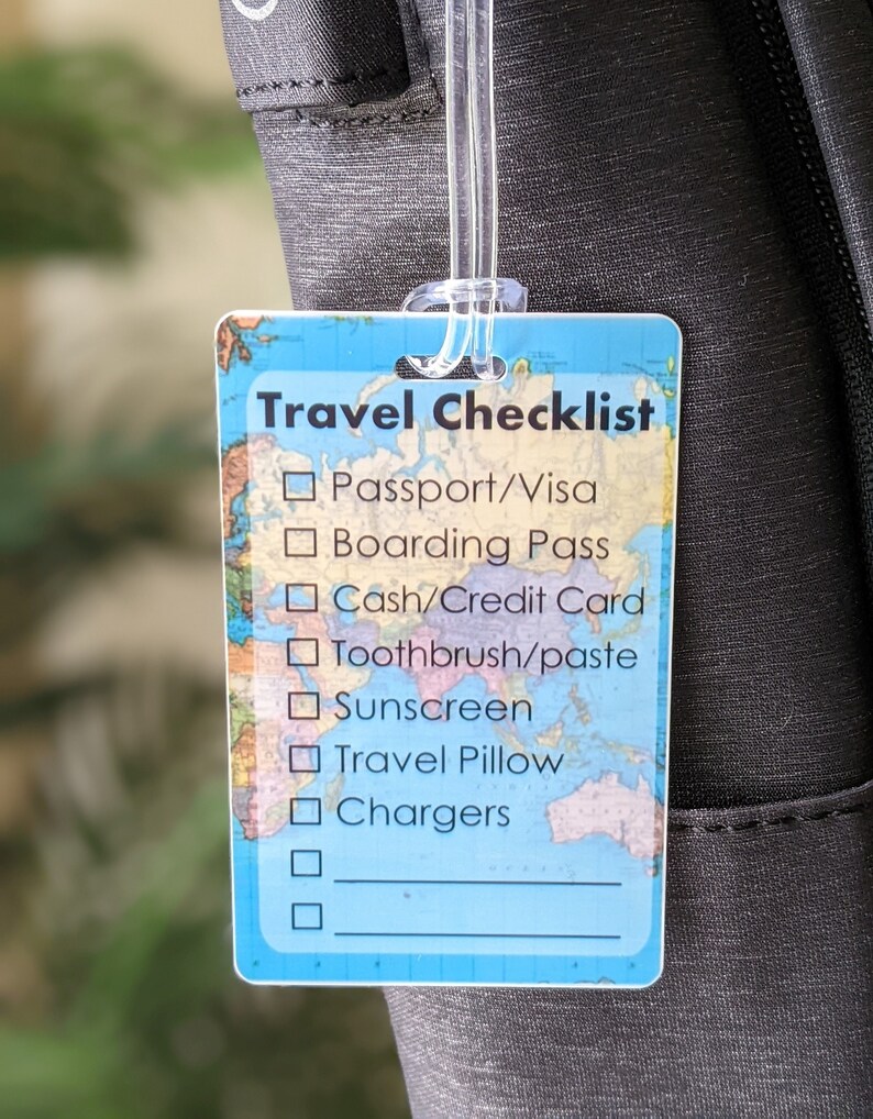 Op de afbeelding: Een rechthoekig reislabel met een doorzichtige plastic lus bevestigd aan een zwarte tas. Het label heeft een blauwe achtergrond met een wereldkaartontwerp en de woorden "Travel Checklist". De checklist bevat items zoals paspoort, boarding pass en opladers.