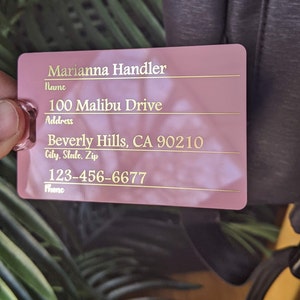 May include: Pink luggage tag with gold lettering. The tag has fields for name, address, city, state, zip, and phone number. The information on the tag reads: Marianna Handler, 100 Malibu Drive, Beverly Hills, CA 90210, 123-456-6677.