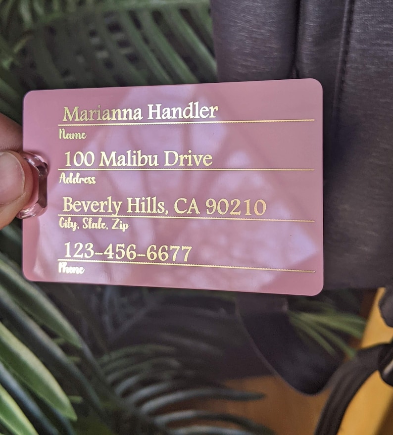 May include: Pink luggage tag with gold lettering. The tag has fields for name, address, city, state, zip, and phone number. The information on the tag reads: Marianna Handler, 100 Malibu Drive, Beverly Hills, CA 90210, 123-456-6677.