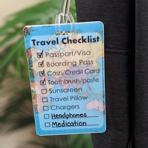 Op de afbeelding: Een rechthoekig reischecklistlabel met een wereldkaartontwerp. Het label heeft een doorzichtige plastic lus. De checklist bevat items zoals paspoort, instapkaart en koptelefoon.