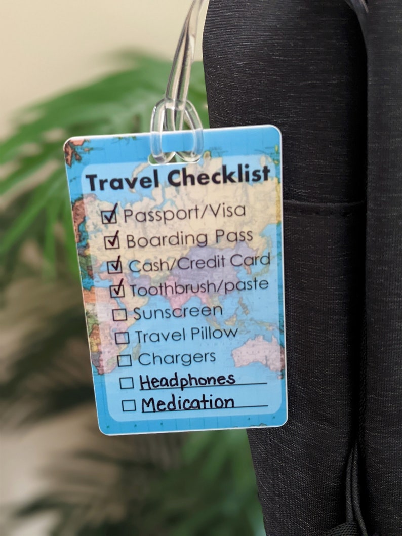 Op de afbeelding: Een rechthoekig reischecklistlabel met een wereldkaartontwerp. Het label heeft een doorzichtige plastic lus. De checklist bevat items zoals paspoort, instapkaart en koptelefoon.