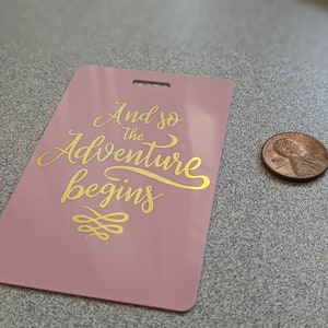 May include: A pink luggage tag with gold lettering that reads "And so the adventure begins" with a gold decorative design below the text.