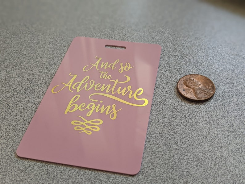 May include: A pink luggage tag with gold lettering that reads "And so the adventure begins" with a gold decorative design below the text.