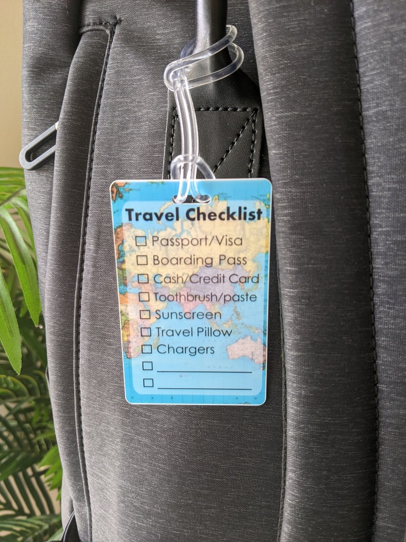 Op de afbeelding: Een rechthoekig reischecklistlabel met een wereldkaartontwerp en een doorzichtige plastic lus. Het label bevat reisbenodigdheden zoals paspoort, instapkaart en opladers. Het label is bevestigd aan een grijze rugzak.