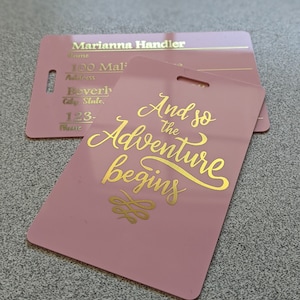 May include: Pink luggage tag with gold lettering that reads "And so the adventure begins".