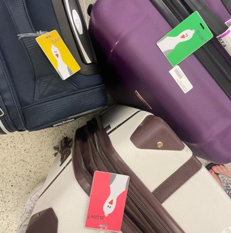 May include: Three suitcases with luggage tags featuring a cartoon goose and the text "mine?" in different colors. The suitcases are purple, white, and navy blue.