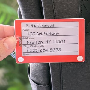 May include: Red luggage tag with white text. The tag has fields for name, address, city, state, zip, and phone number. The text on the tag reads: "E. Sketcherson", "100 Art Parkway", "New York, NY 14301", "(555) 234-5678".