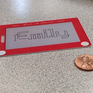May include: Red Etch A Sketch toy with a white screen and a white knob on each side. The screen has the name "Emily" drawn on it in black.
