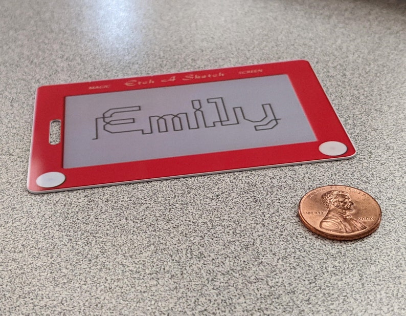 May include: Red Etch A Sketch toy with a white screen and a white knob on each side. The screen has the name "Emily" drawn on it in black.