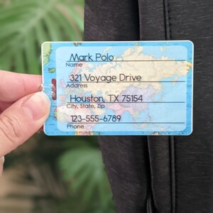 Op de afbeelding: Een rechthoekig bagagelabel met een wereldkaartontwerp. Het label toont de naam "Mark Polo", adres "321 Voyage Drive, Houston, TX 75154" en telefoonnummer "123-555-6789". Het label is tegen een donkere achtergrond.