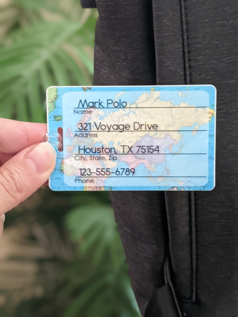 Op de afbeelding: Een rechthoekig bagagelabel met een wereldkaartontwerp. Het label toont de naam "Mark Polo", adres "321 Voyage Drive, Houston, TX 75154" en telefoonnummer "123-555-6789". Het label is tegen een donkere achtergrond.