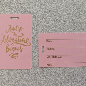 May include: Two pink luggage tags with gold lettering. The tag on the left says "And so the adventure begins" with a decorative swirl. The tag on the right has blank lines for name, address, city, state, zip, and phone.