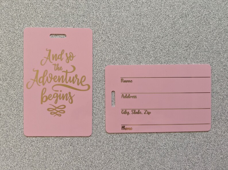 May include: Two pink luggage tags with gold lettering. The tag on the left says "And so the adventure begins" with a decorative swirl. The tag on the right has blank lines for name, address, city, state, zip, and phone.