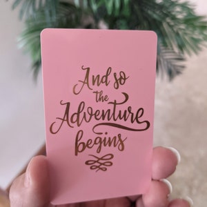May include: Pink card with gold lettering that reads "And so the adventure begins" with a decorative flourish.