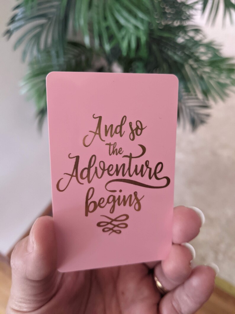 May include: Pink card with gold lettering that reads "And so the adventure begins" with a decorative flourish.