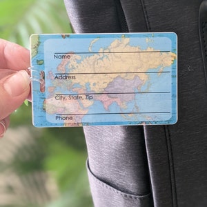 Op de afbeelding: Een rechthoekig bagagelabel met een wereldkaartontwerp. Het label heeft een doorzichtige plastic lus voor bevestiging en bevat velden voor naam, adres, stad, postcode en telefoon. De achtergrond is donkergrijs.