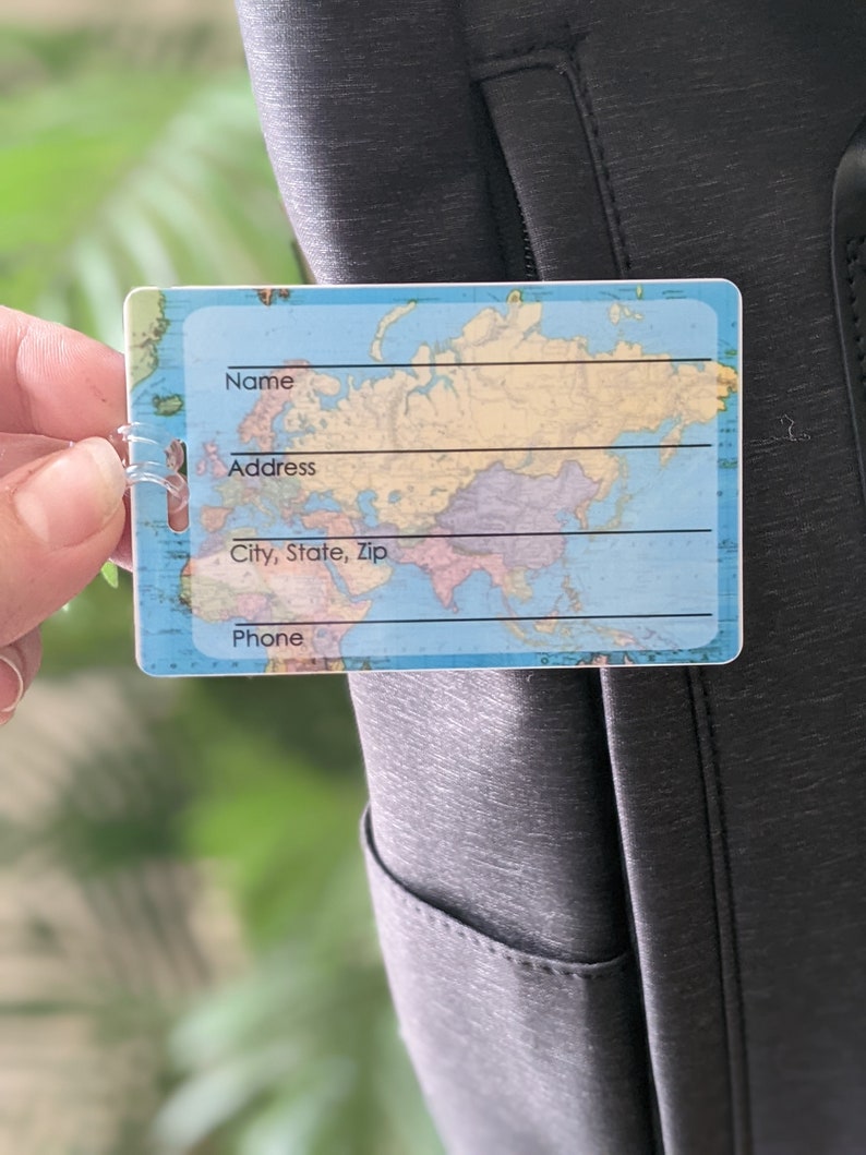 Op de afbeelding: Een rechthoekig bagagelabel met een wereldkaartontwerp. Het label heeft een doorzichtige plastic lus voor bevestiging en bevat velden voor naam, adres, stad, postcode en telefoon. De achtergrond is donkergrijs.