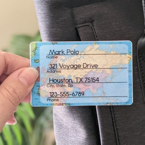 Op de afbeelding: Een rechthoekig bagagelabel met een wereldkaartontwerp. Het label toont de naam "Mark Polo", adres "321 Voyage Drive, Houston, TX 75154" en telefoonnummer "123-555-6789". Het label wordt tegen een donkergrijze tas gehouden.