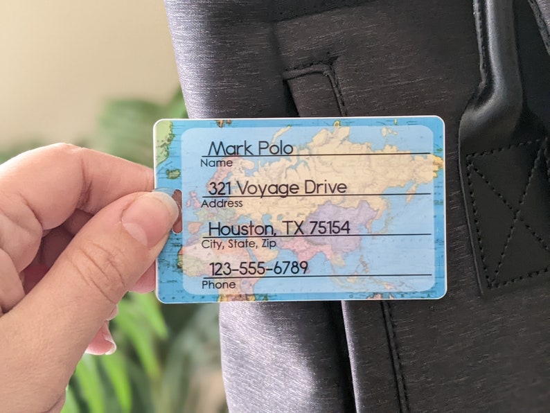 Op de afbeelding: Een rechthoekig bagagelabel met een wereldkaartontwerp. Het label toont de naam "Mark Polo", adres "321 Voyage Drive, Houston, TX 75154" en telefoonnummer "123-555-6789". Het label wordt tegen een donkergrijze tas gehouden.