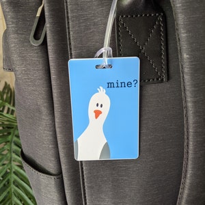 May include: A blue luggage tag with a white cartoon bird and the text "mine?" printed on it.
