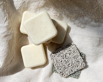 May include: Four white square-shaped soap bars stacked on top of each other on a white fabric background. A gray and white patterned soap saver is in the lower right corner of the image.