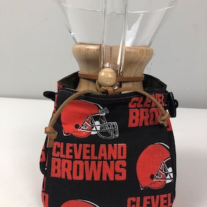 May include: A black fabric bag with a drawstring closure and a brown leather strap. The bag has a repeating pattern of orange and white Cleveland Browns logos. The bag is holding a clear glass pour-over coffee maker with a wooden base.