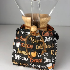 May include: A black fabric bag with a brown leather drawstring closure. The bag is printed with various coffee drink names in white and orange text. A glass and wood pour-over coffee maker is inside the bag.