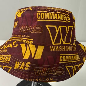 May include: A burgundy and gold bucket hat featuring the text "COMMANDERS" and "WAS WASHINGTON" in a repeating pattern. The hat has a wide brim and a round crown. The hat is made of a soft fabric.