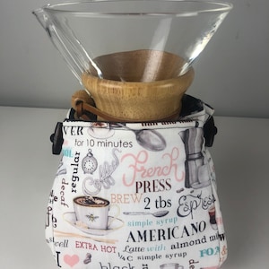 May include: A white fabric coffee bag with a black strap and a glass coffee filter on top. The bag has a coffee themed print with text that says "French Press Brew 2 tbs simple syrup Americano Latte with almond milk 1/4C simple syrup".