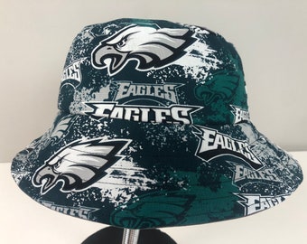 May include: A bucket hat featuring a repeating pattern of the Philadelphia Eagles logo and the word "EAGLES" in white and gray on a dark green background. The hat has a wide brim and a rounded crown.