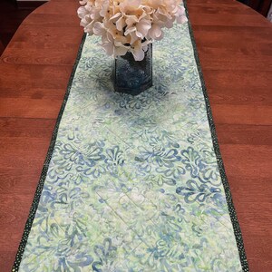 Pu&ograve; includere: Un runner da tavola trapuntato rettangolare con un motivo in stile batik nei toni del verde chiaro, blu e bianco. Il runner presenta un bordo verde scuro con piccoli puntini bianchi e cuciture diagonali a macchina sulla superficie.