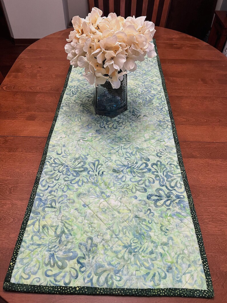 Pu&ograve; includere: Un runner da tavola trapuntato rettangolare con un motivo in stile batik nei toni del verde chiaro, blu e bianco. Il runner presenta un bordo verde scuro con piccoli puntini bianchi e cuciture diagonali a macchina sulla superficie.