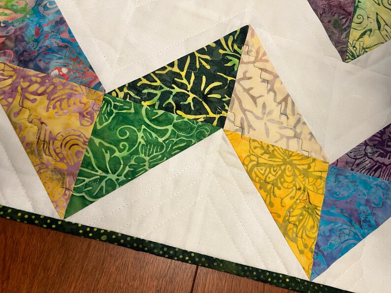 Pu&ograve; includere: Un pezzo di tessuto trapuntato caratterizzato da un motivo geometrico di forme triangolari in stampe batik gialle, verdi, blu e viola, intervallate da sezioni di tessuto bianco tinta unita. I bordi sono rifiniti con un bordo verde scuro a pois.