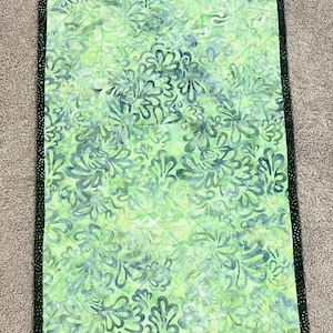 Pu&ograve; includere: Un runner da tavola rettangolare con un motivo floreale in stile batik nei toni del verde chiaro e scuro. Il tessuto &egrave; trapuntato con linee di cucitura diagonali e rifinito con un bordo verde scuro caratterizzato da piccole macchie chiare.